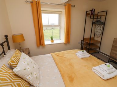 A bedroom with a bed with yellow and floral pillows a yellow blanket and folded towels on it a window with yellow curtains a bedside table with a yellow lamp and a wooden shelving unit at Henhill Lodge in Chathill near Seahouses