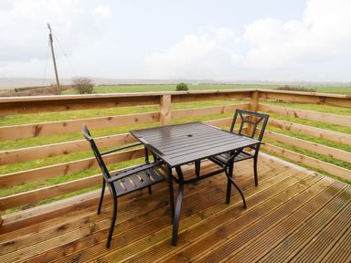 A wooden deck with a black metal table and two chairs overlooking a green field at Henhill Lodge in Chathill near Seahouses