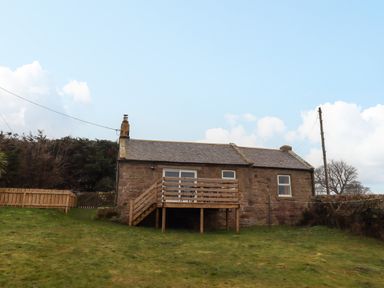A stone cottage with a wooden deck and stairs on a grassy lawn at Henhill Lodge in Chathill near Seahouses