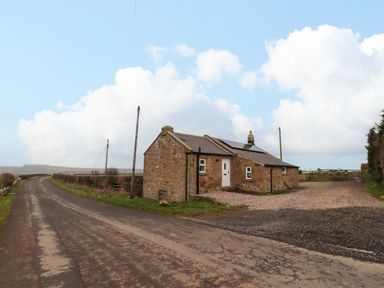 A stone cottage with a white door and solar panels on the roof beside a country road at Henhill Lodge in Chathill near Seahouses