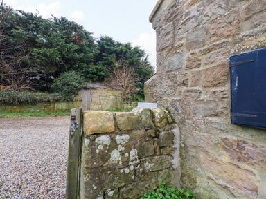 A stone gate and wall next to a stone building with a blue shutter and shrubbery at Henhill Lodge in Chathill near Seahouses