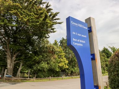 A blue sign for Green Hill Farm holiday village among trees and a road at 7 Wild Haven in Landford