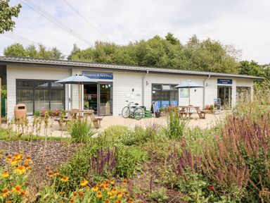 An outdoor area with two umbrella-shaded picnic tables in front of a reception and park shop building surrounded by plants and trees at 7 Wild Haven in Landford