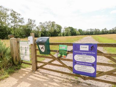 A wooden gate with dog waste bin and signage opening to a gravel path in a field at 7 Wild Haven in Landford