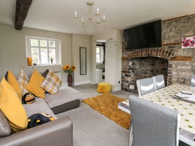 A living room with a sofa and yellow pillows a dining table with chairs a wall clock and a television at Yewtree Cottage in Defynog near Sennybridge
