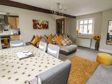 A living room with a dining table covered with a bee-patterned tablecloth chairs a sofa with yellow and plaid cushions a yellow rug and a yellow pouf at Yewtree Cottage in Defynog near Sennybridge