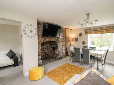 A living room with a stone fireplace and mounted TV a dining table with six chairs and a window with plaid curtains at Yewtree Cottage in Defynog near Sennybridge
