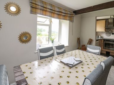 A dining area with bee patterned tablecloth and six chairs by a window with plaid curtains and sunburst mirrors at Yewtree Cottage in Defynog near Sennybridge