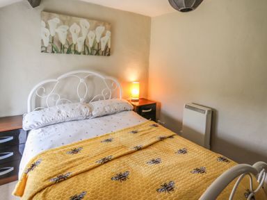 A bedroom with a white metal bed frame a yellow blanket with bee patterns a bedside table with a lamp and a painting of white flowers on the wall at Yewtree Cottage in Defynog near Sennybridge