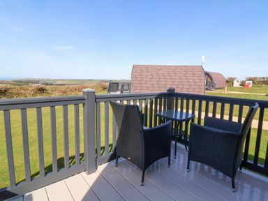 An outdoor deck with two black chairs and a small table overlooking grassy area and brown roofed accommodation at Owl's Nook in Kilkhampton