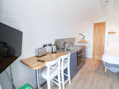 A small kitchen area with two white chairs a wooden countertop a kettle toaster and stove next to a bed and door at Owls Nook in Kilkhampton