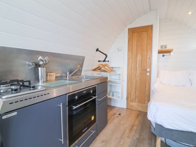 A compact kitchen area with stovetop oven sink and utensils next to a wooden door and a bed in a small curved room at Owls Nook in Kilkhampton