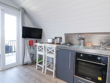 A kitchenette with an oven, stove, sink, toaster, kettle, two white bar stools, a wall-mounted TV, and a door leading to an outdoor deck at Owl's Nook in Kilkhampton