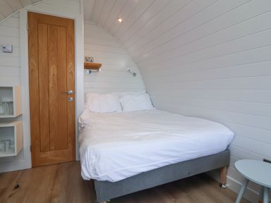 A bedroom with a double bed white bedding wooden door small round table and built in shelves with cups at Owls Nook in Kilkhampton