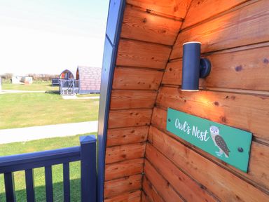 Wooden cabin corner with a black wall light and an Owls Nest sign on the wall with other cabins and grassy field in the background at Owls Nook in Kilkhampton