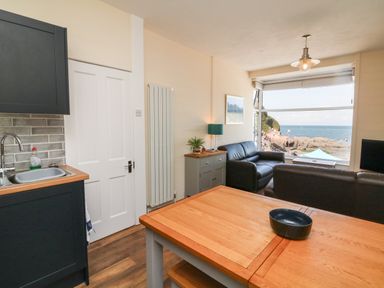 A living area with a wooden table a black bowl two black sofas a sideboard with a lamp and plant a kitchen sink and cabinets and a window showing a coastal view at At the Bay Apartment in Hele Bay Ilfracombe
