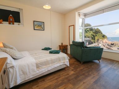 A bedroom with a bed a green sofa wooden side table a mirror and a large window overlooking a coastal area at At the Bay Apartment in Hele Bay Ilfracombe