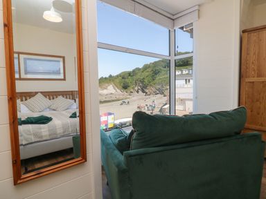 A room with a large window overlooking a beach a green sofa a wooden wardrobe and a mirror reflecting a bed at At the Bay Apartment in Hele Bay Ilfracombe