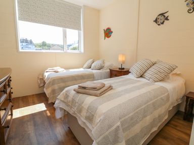 A bedroom with two single beds a nightstand with a lamp a dresser and windows at At the Bay Apartment in Hele Bay Ilfracombe