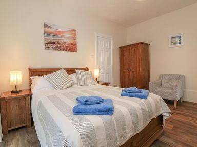 A bedroom with a double bed with blue towels on it two bedside tables with lamps a wooden wardrobe and a patterned armchair at At the Bay Apartment in Hele Bay Ilfracombe