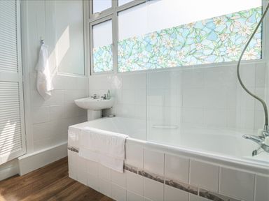 A bathroom with a bathtub a sink a towel hanging on a hook and a window with floral stained glass at At the Bay Apartment in Hele Bay Ilfracombe
