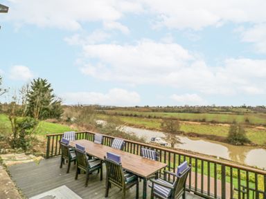 An outdoor deck with a wooden table and chairs overlooking a river and grassy fields at Riverside Manor in Evesham
