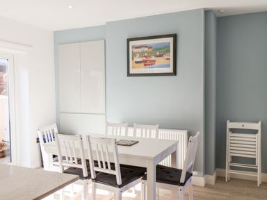 A dining area with a white table and six white chairs with black cushions a wall cabinet and a framed picture of boats on a blue wall at Whitby View in Whitby