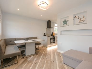 A dining area with grey bench seating and a white table next to a kitchen with stainless steel appliances and wooden flooring at Number 9 in California Norfolk