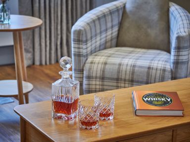 A wooden table with a crystal decanter and two glasses containing whisky and a book titled World Whisky next to a plaid armchair at The Filly in Humshaugh