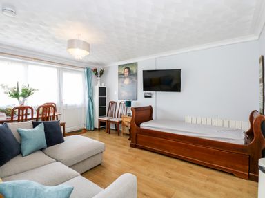 A living room with a grey sofa, wooden daybed, dining table with chairs, wall-mounted TV and a painting at 63 The Waterside Holiday Park in Corton