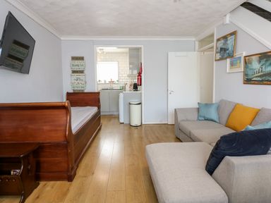 A living room with a wooden daybed a gray sofa with colored cushions wall art and a kitchen visible through a doorway at 63 The Waterside Holiday Park in Corton