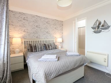 A bedroom with a double bed and bedside tables with lamps a door leading to a bathroom and a wall decoration of sailboats at 63 The Waterside Holiday Park in Corton