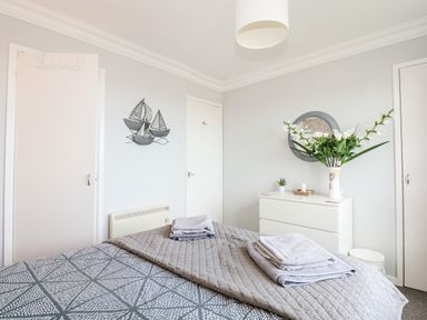 A bedroom with a bed covered in gray bedding and towels on top a white dresser with flowers and a round mirror on the wall at 63 The Waterside Holiday Park in Corton