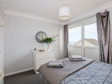 A bedroom with a bed, folded towels, a dresser with flowers, a round wall mirror, and large windows with curtains at 63 The Waterside Holiday Park in Corton