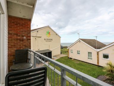 A balcony with two chairs and a small round table overlooking grass and buildings near the sea at 63 The Waterside Holiday Park in Corton