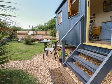 An outdoor garden area with a small table and two chairs next to stairs leading to a blue cabin with an open door at Dot May Porteath near Polzeath