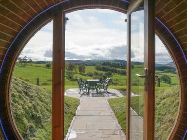 A round doorway opens to a stone patio with a table and two benches overlooking a green landscape at Badgers View in Rhayader