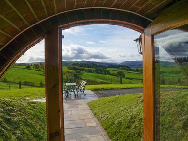 A view through an arched wooden door showing a stone pathway leading to a metal table and benches on a patio with green fields and hills in the background at Badgers View in Rhayader