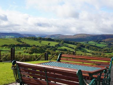 A wooden bench and metal table on grass overlooking green hills and trees at Badgers View in Rhayader