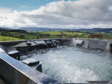 A hot tub with bubbling water overlooking green fields and hills with a metal bench and table set in the background at Badgers View in Rhayader