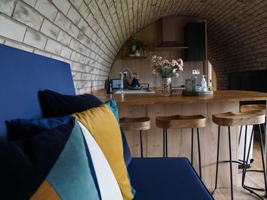 A kitchen with a wooden island and stools with a blue sofa and colorful cushions in a space with a curved brick ceiling at Badgers View in Rhayader