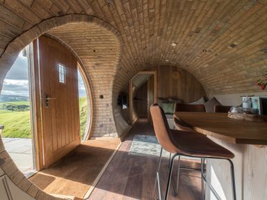 An interior of a curved brick structure with a wooden door open to a grassy outside area showing a table and chairs inside at Hares View in Rhayader