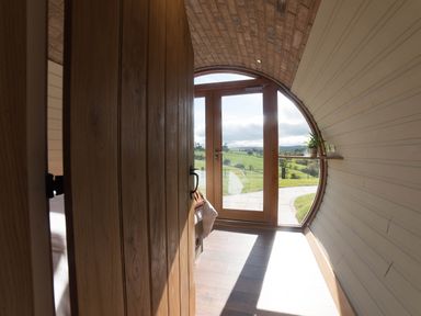 A wooden door opens to a curved room with wood paneling and a glass door showing green fields at Hares View in Rhayader