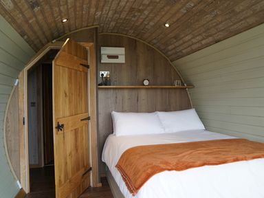 A bedroom with a bed with white bedding and an orange blanket an open wooden door and a wooden shelf with a clock and lamp at Hares View in Rhayader