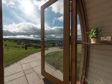 A glass door open to a paved patio and green fields with a plant on a wooden shelf inside at Hares View in Rhayader