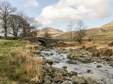 A stone bridge over a rocky stream with trees and hills in the background at Gosling Cottage in Kendal
