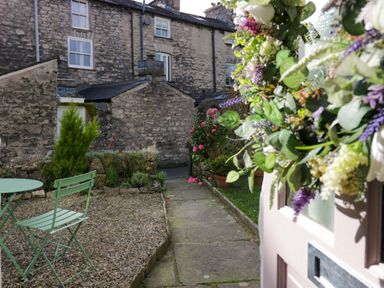 A small garden area with a green metal table and chair near stone buildings and a floral wreath on a door at Gosling Cottage in Kendal