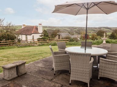 An outdoor patio with a round glass table and wicker chairs under a large umbrella with a grassy yard and hills in the background at Fountain Folly in Moylgrove near St Dogmaels