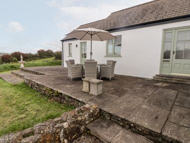 An outdoor stone patio with a table and chairs under an umbrella next to a white building at Fountain Folly in Moylgrove near St Dogmaels