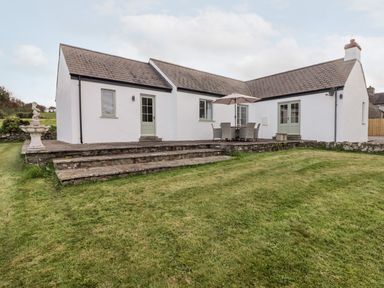 A white house with stone steps leading to a patio with table chairs and umbrella and a green lawn at Fountain Folly in Moylgrove near St Dogmaels